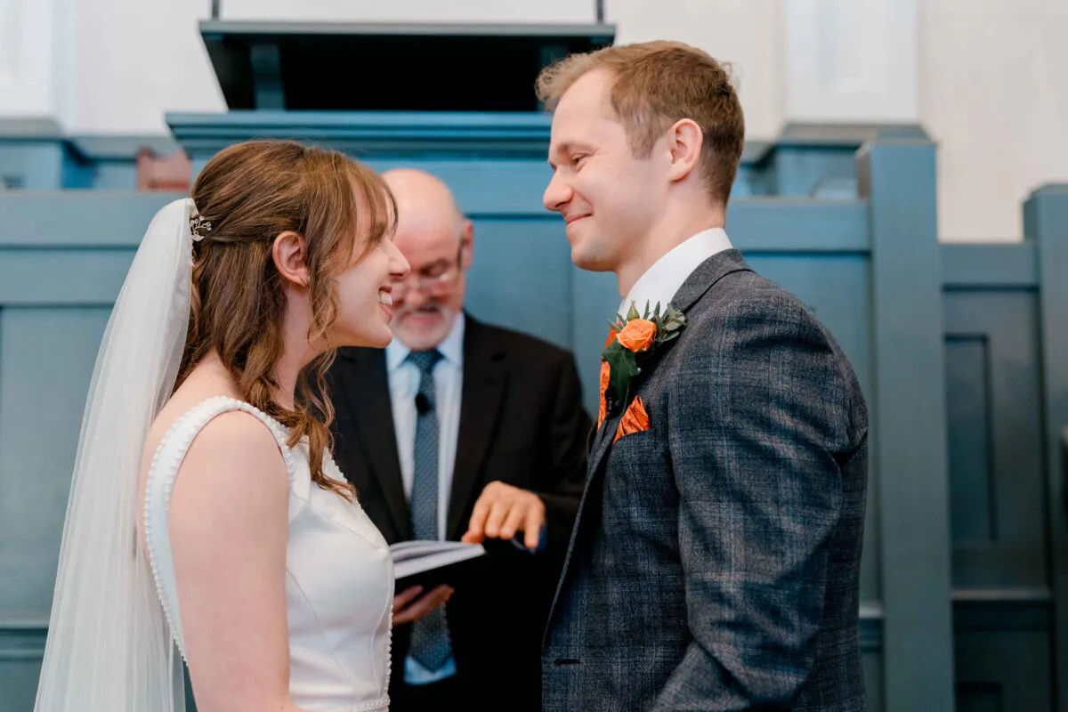 A smiling bride and groom look lovingly at each other during their indoor wedding ceremony while an officiant conducts the service in the background.