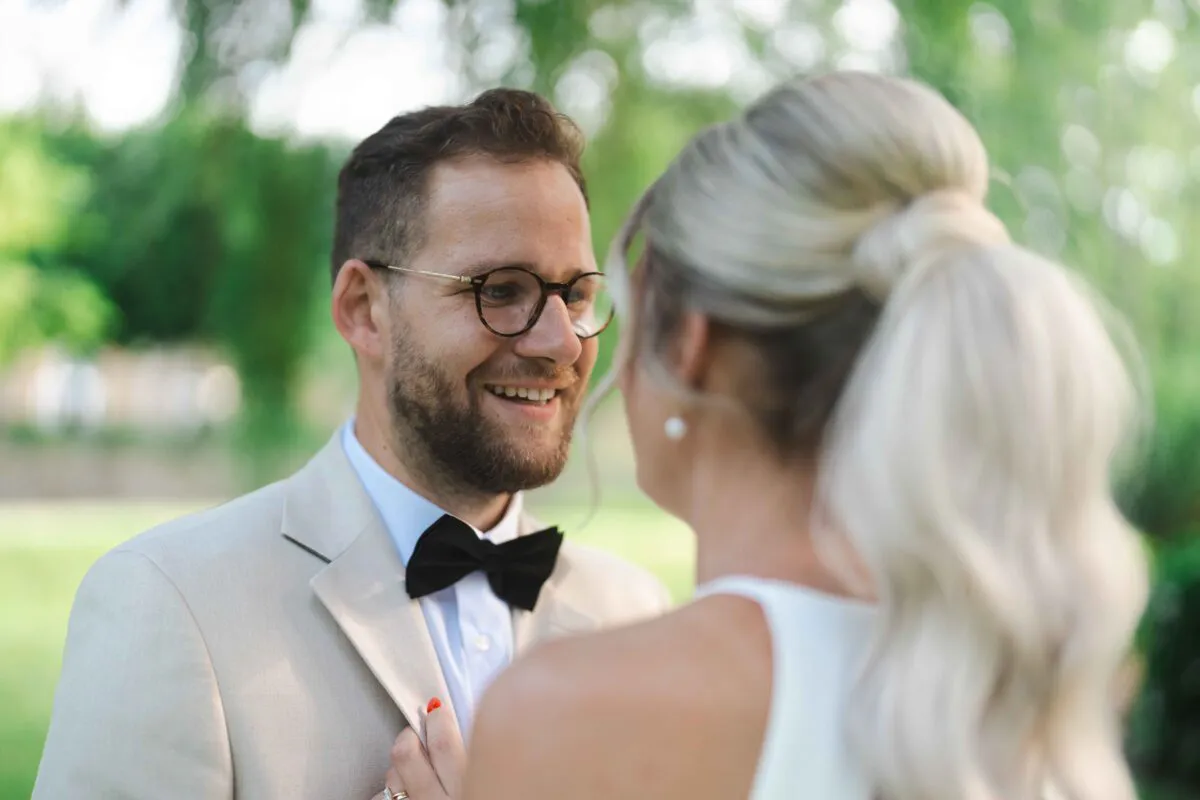 A smiling groom in a beige suit and black bow tie looks lovingly at his bride outdoors during their wedding portrait session.