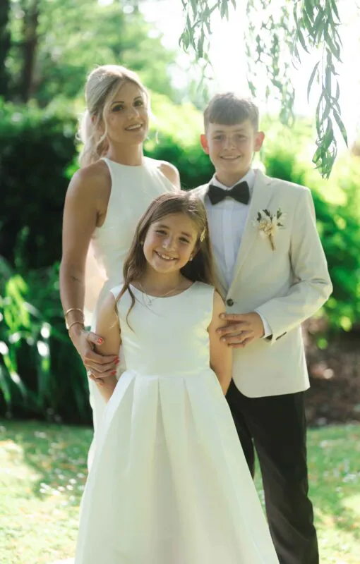 A smiling family group consisting of a woman in a white halterneck dress, a young girl in a white dress, and a young boy in a cream suit jacket with a bow tie stand outdoors in a sunlit garden.