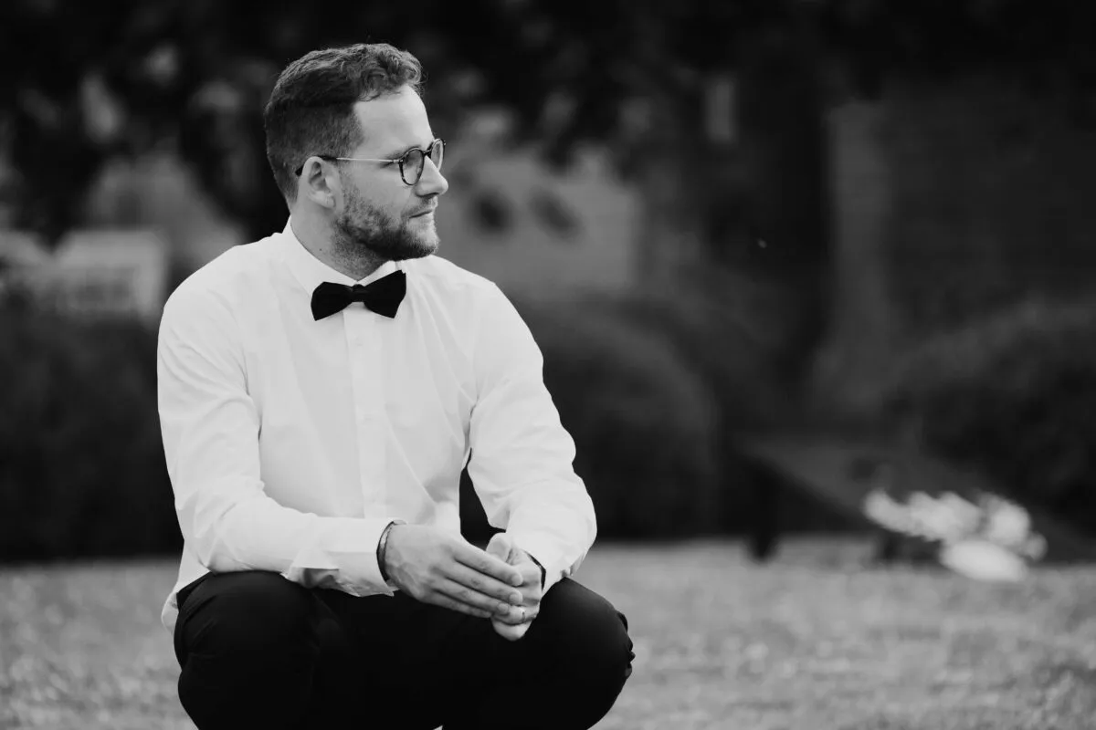 A stylish groom wearing a crisp white shirt and black bow tie kneels outdoors in a classic black and white portrait.