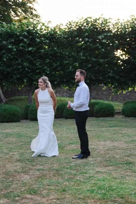 A joyful bride in a modern halter neck gown and groom share a happy laugh together outdoors in a manicured garden setting.