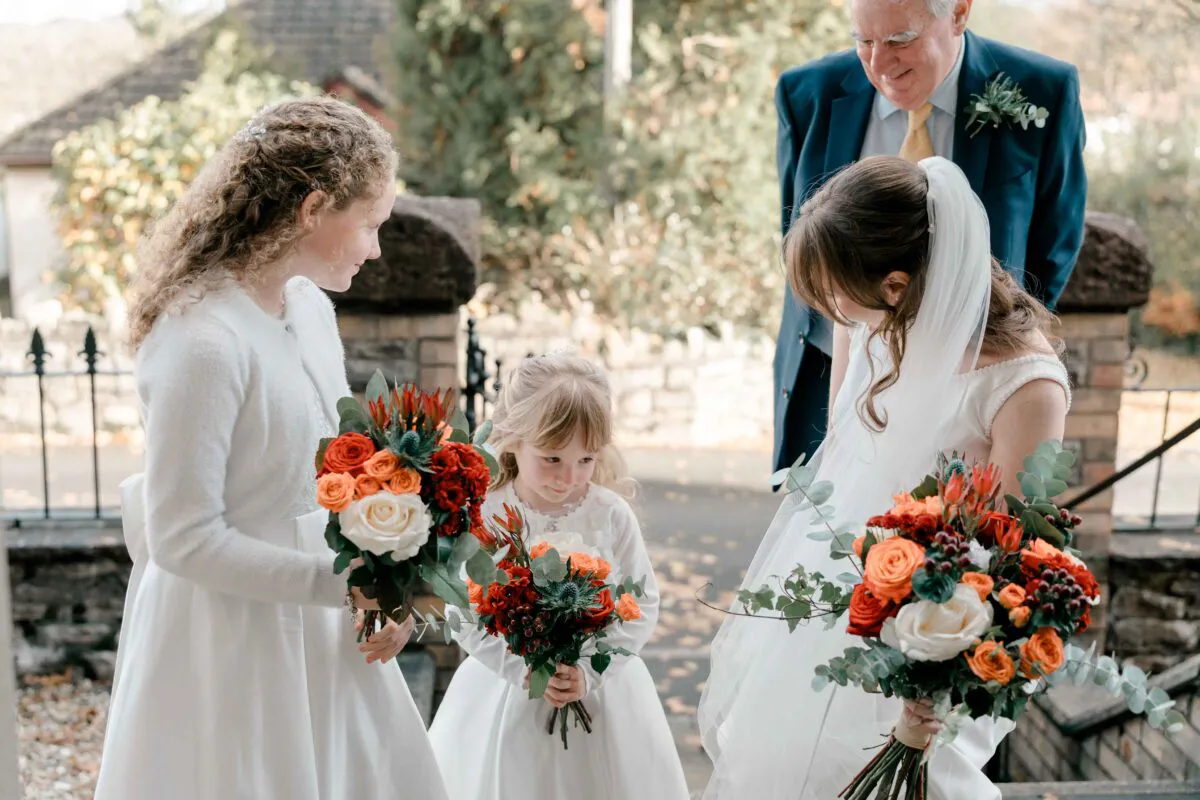 A young bride holding a vibrant autumn-toned bouquet stands alongside two flower girls and an older male relative outside a stone venue.