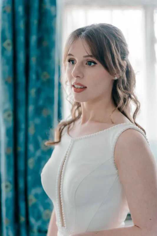 A stunning bridal portrait of a bride getting ready, featuring a half-up half-down hairstyle with soft waves, curtain bangs, pearl earrings, and a crepe wedding dress with pearl detailing along the neckline.