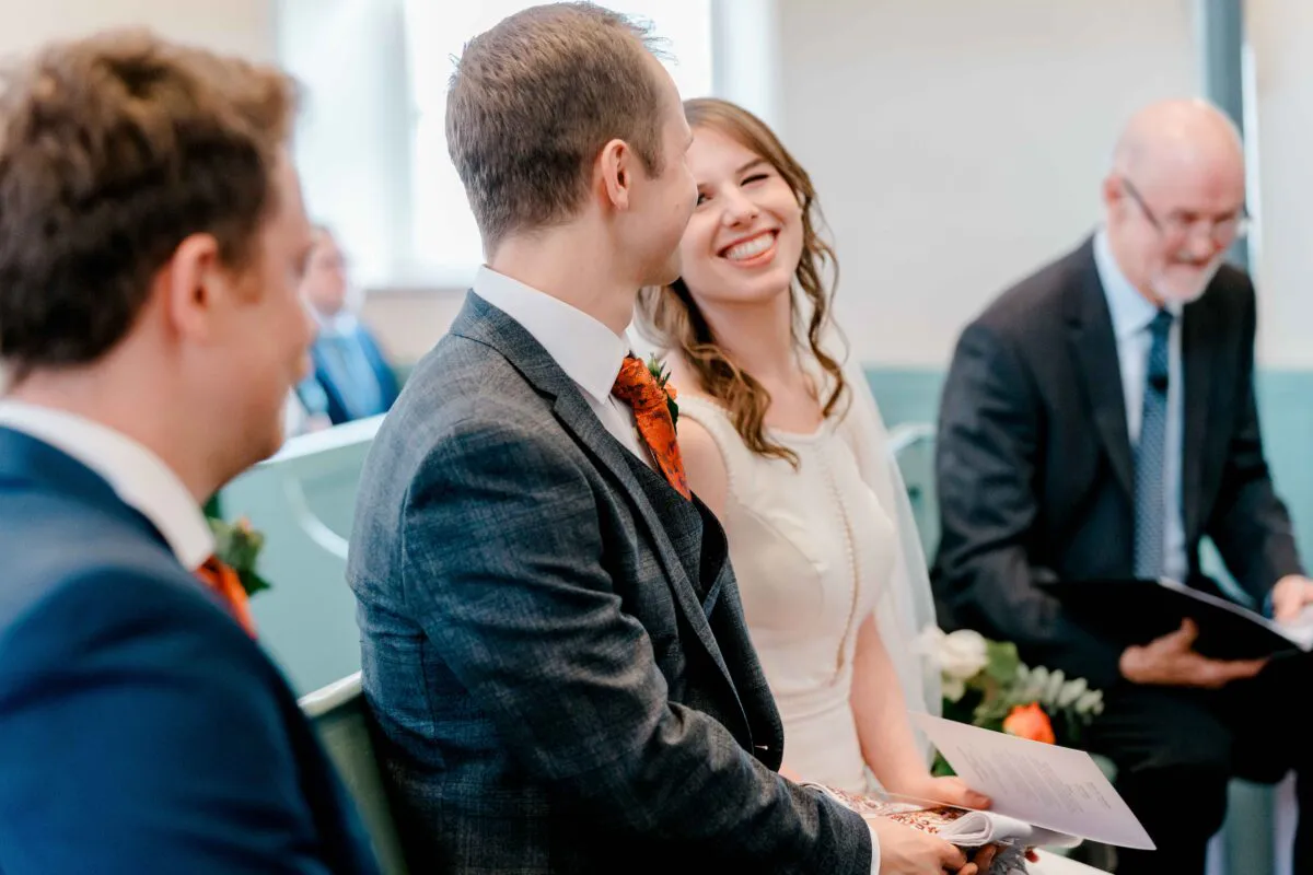 A smiling bride and groom sit together during their wedding ceremony, sharing a joyful moment of laughter with family and guests nearby.