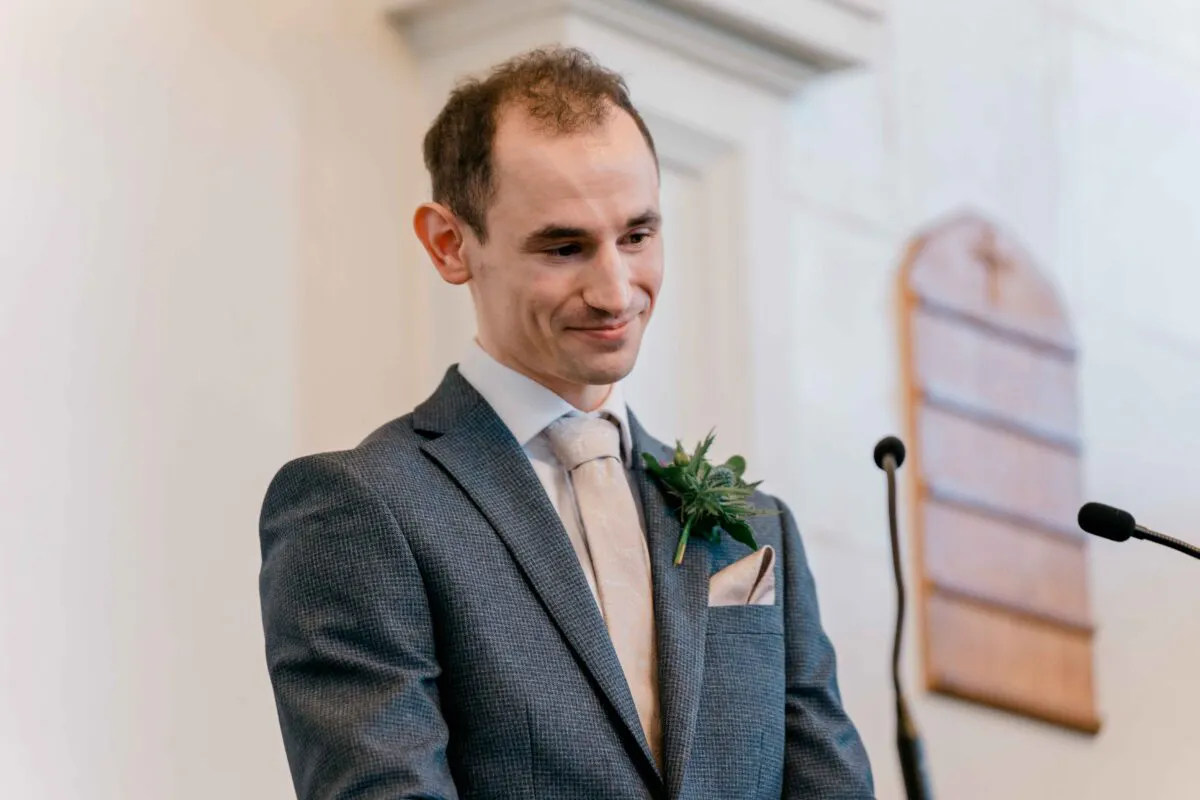 A smiling groom wearing a grey textured suit and pale tie stands at the front of a church during the wedding ceremony.
