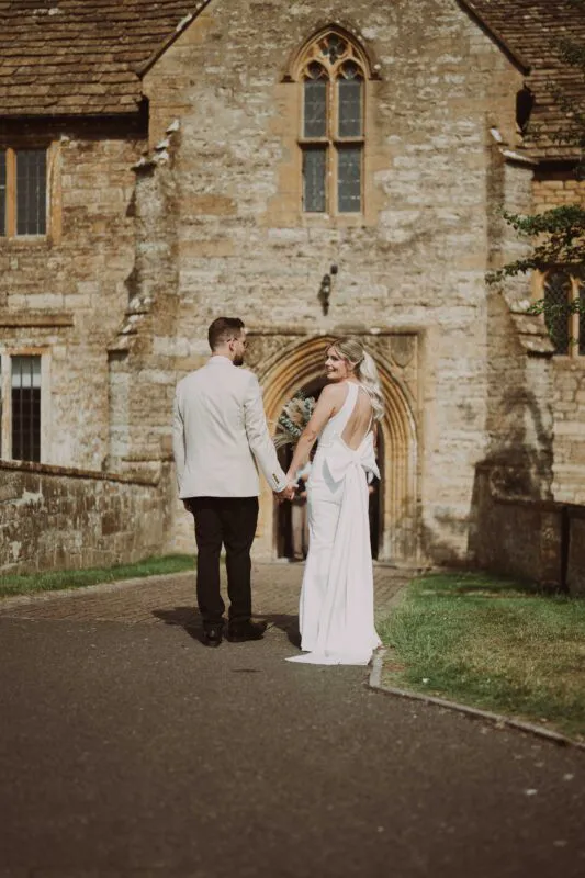 A bride and groom walk hand-in-hand along a paved pathway outside a historic stone manor house, with the bride looking back over her shoulder with a smile.