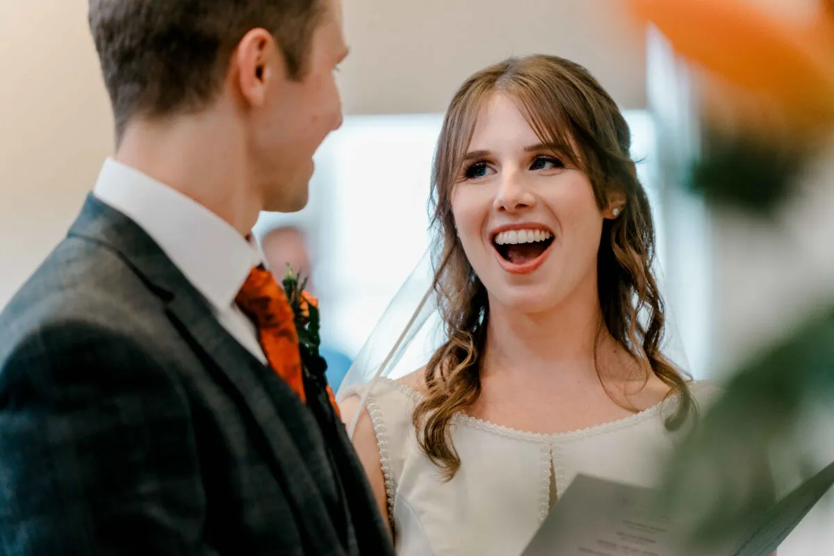 A joyful bride laughs happily while looking at her groom during the wedding ceremony, wearing a pearl-trimmed gown and veil.