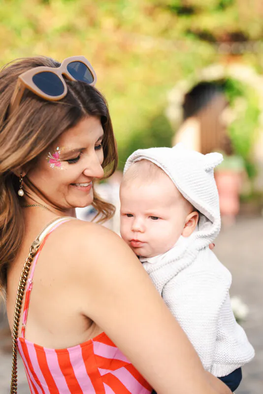 A guest at an outdoor wedding holds a baby wearing a white hooded knit jumper while smiling outdoors in natural light.