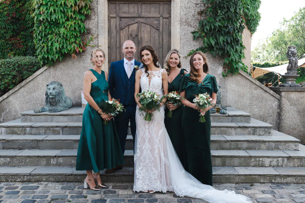 A smiling bride and groom pose on stone steps outside a wooden doorway with three bridesmaids wearing dark green dresses and holding bouquets.