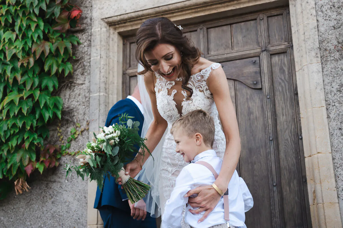 A laughing bride holding a bouquet shares a joyful moment with a young page boy outside a stone church entrance covered in ivy.