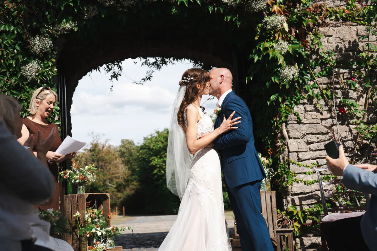 A newlywed couple share their first kiss beneath an ivy-covered stone archway during an outdoor ceremony while guests applaud.