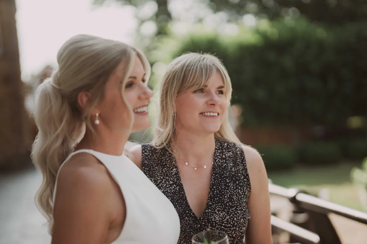 Two smiling women standing outdoors at a wedding reception, sharing a happy moment together in natural light.