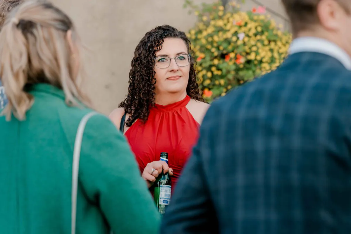 A wedding guest with dark curly hair and glasses wearing a red dress holds a glass bottle while mingling outdoors.