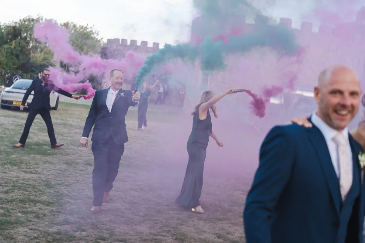 A joyous wedding party and guests celebrate outdoors on a grassy lawn with colourful pink and green smoke bombs going off in front of a castle.