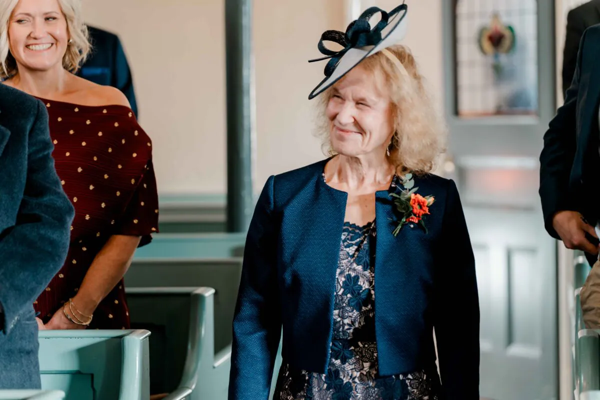 An elderly female guest wearing a navy jacket and a decorative fascinator smiles warmly inside a church during the wedding ceremony.