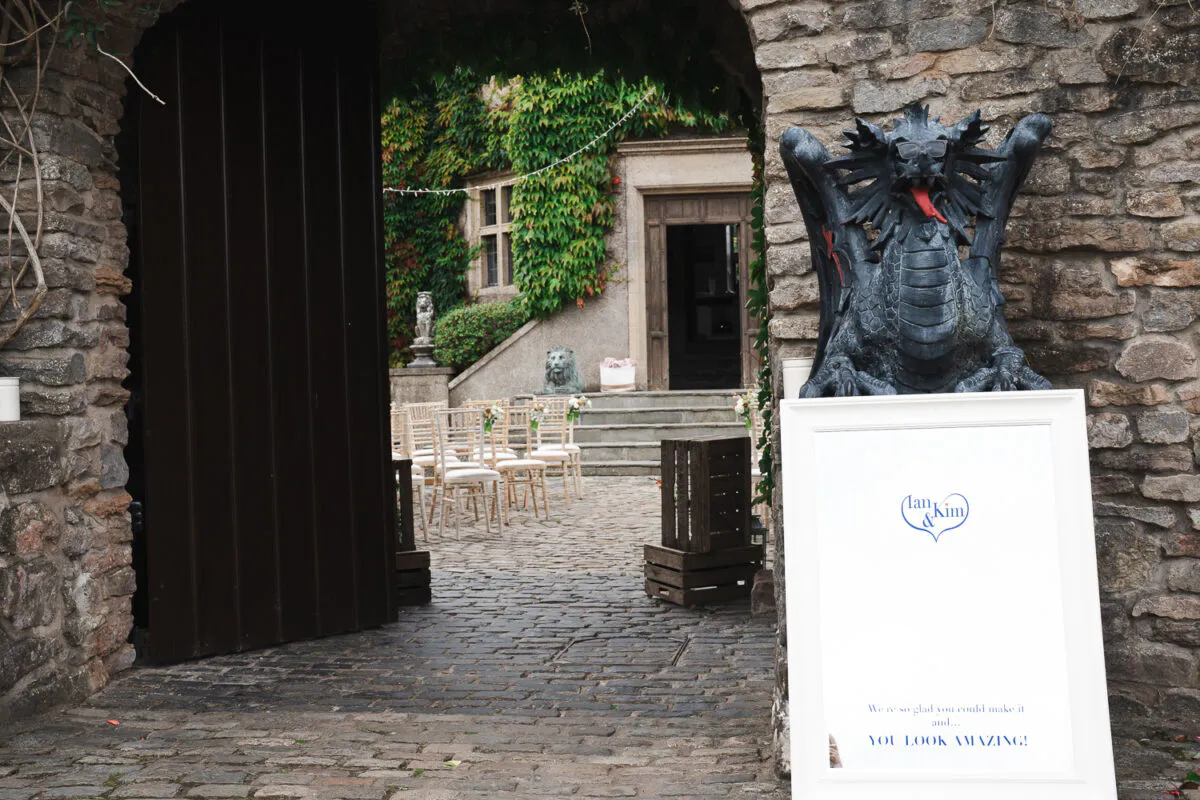 A welcoming wedding sign featuring the couple's names stands outside a historic castle entrance next to a whimsical stone dragon statue.