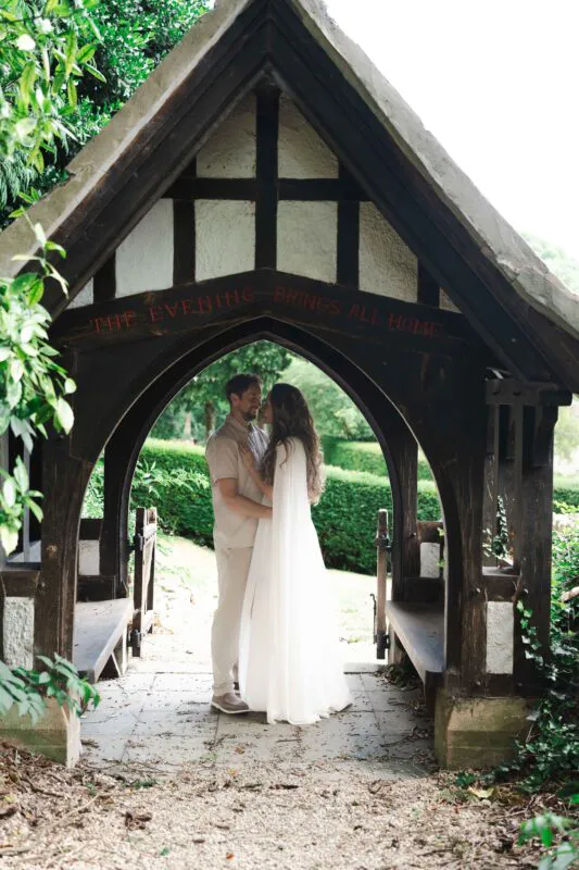 A newlywed couple embrace nose-to-nose beneath a timber and stone lychgate featuring carved lettering reading the evening brings all home.