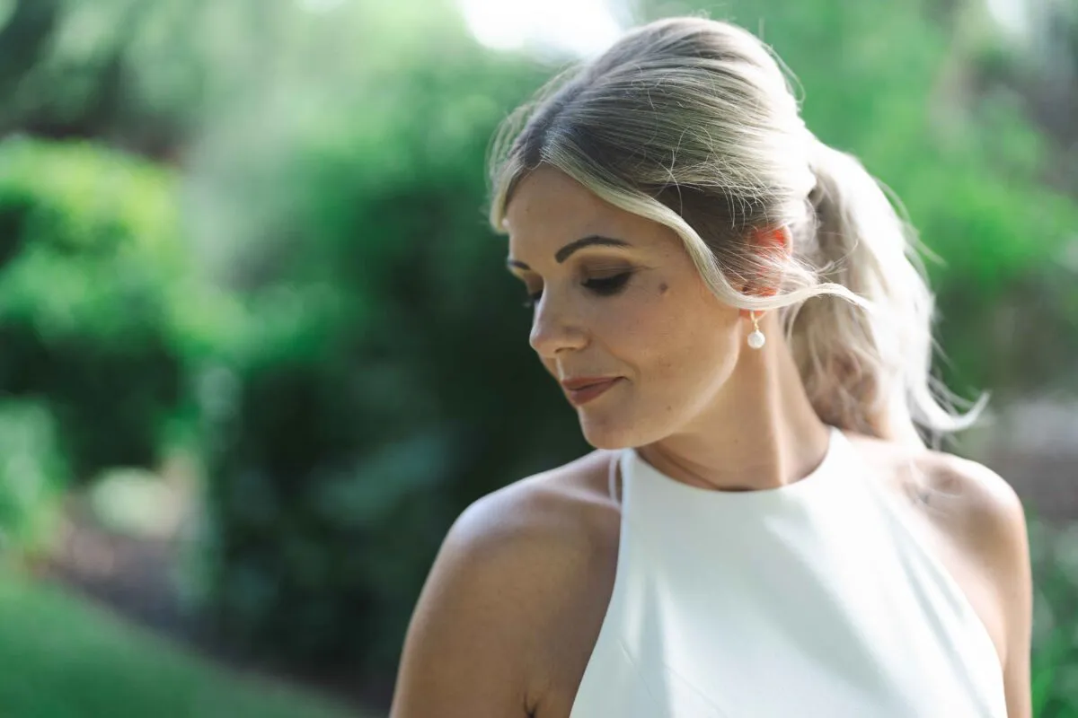 A close-up portrait of a bride wearing a halter neck dress and pearl earrings, with her blonde hair styled in a low ponytail, looking down softly against a lush green background.