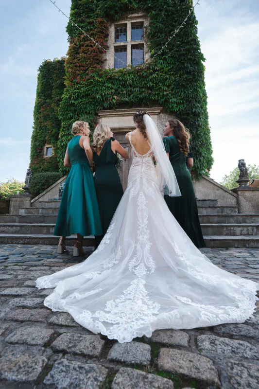 A bride in an intricate lace gown with a long cathedral train stands with her bridesmaids in emerald green dresses in front of an ivy-covered stone tower.