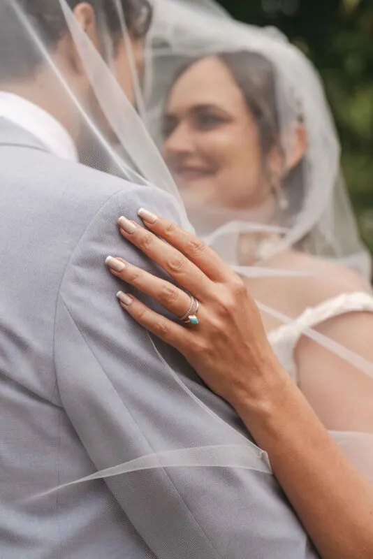A bride rests her hand gently on the groom's grey suited shoulder, with both of them tenderly framed beneath a soft tulle veil.