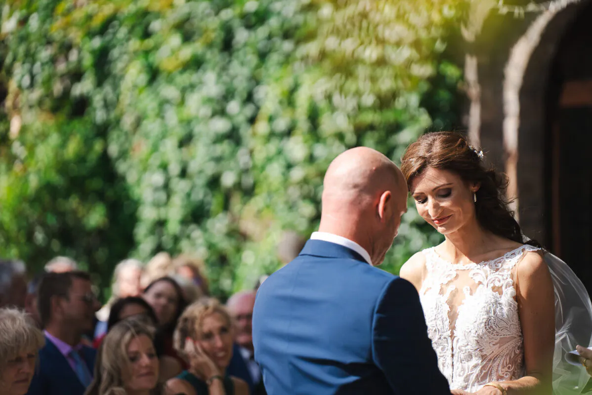 A bride in an illusion neckline lace dress and a groom in a blue suit stand together during an outdoor ceremony against an ivy-covered wall.