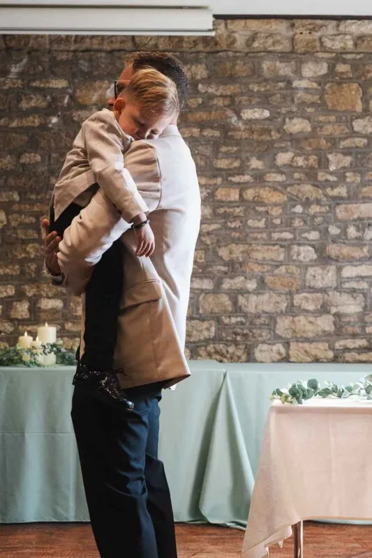 A man in a beige suit lovingly holds a tired page boy resting his head on his shoulder against a rustic stone wall background.
