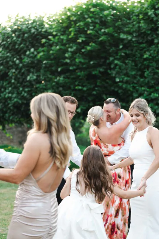 Wedding guests enjoy a joyful outdoor celebration in a lush garden setting as children and adults dance together in the warm sunlight.
