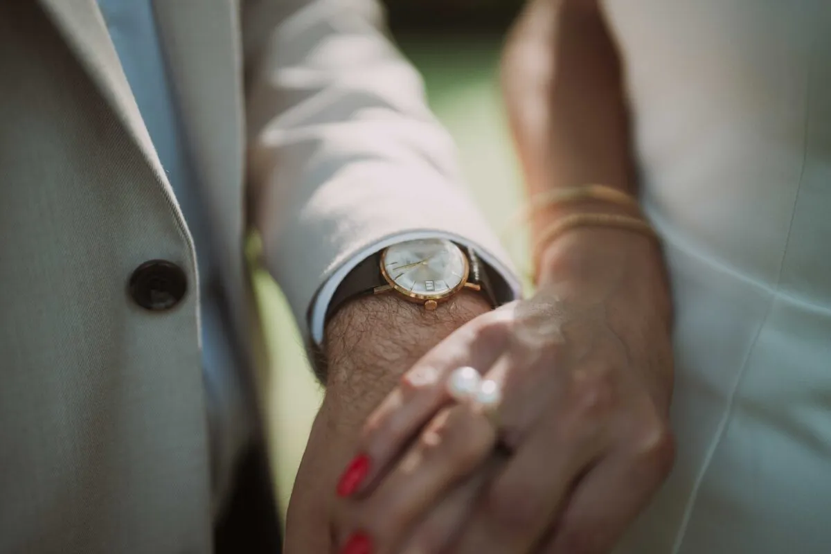 Close-up of a couple holding hands, highlighting a groom's elegant wristwatch and the bride's delicate stacked rings against a neutral suit.