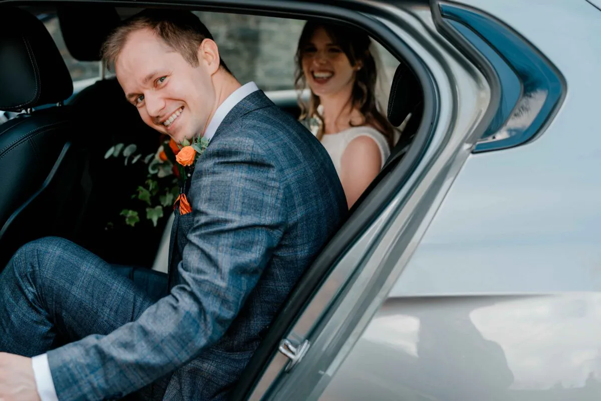 A smiling groom in a checked suit looks back from the backseat of a car while the bride laughs in the background.