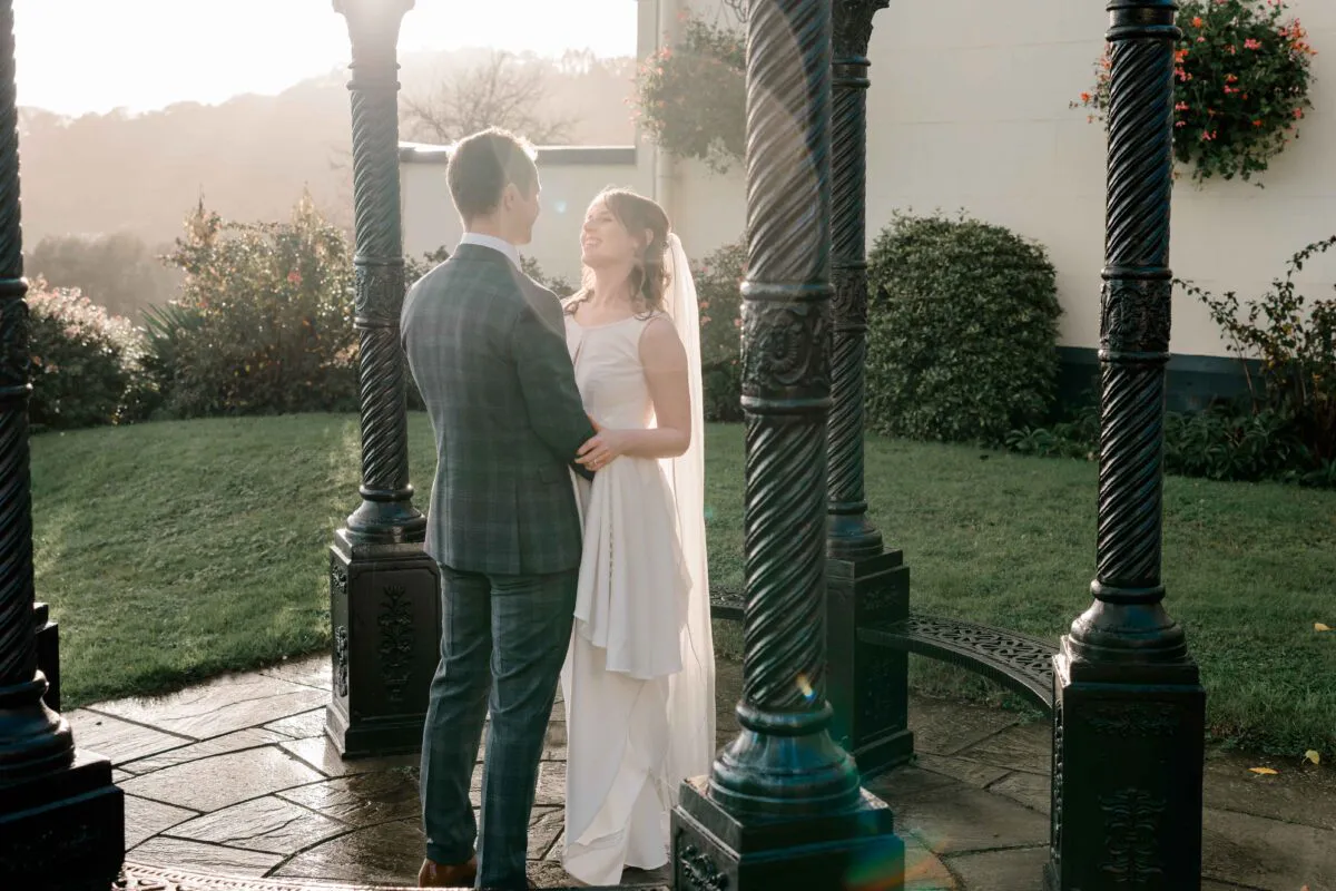 A newlywed couple share a joyful moment standing inside an ornate black metal bandstand during golden hour.