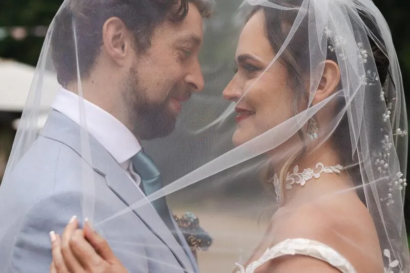 A newlywed couple share a tender moment nose to nose underneath a sheer pearl-embellished veil outdoors.