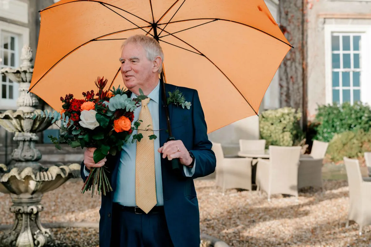 An older gentleman wearing a navy suit and yellow tie stands outdoors holding a vibrant orange umbrella and a colourful bouquet of autumn flowers near a stone fountain.