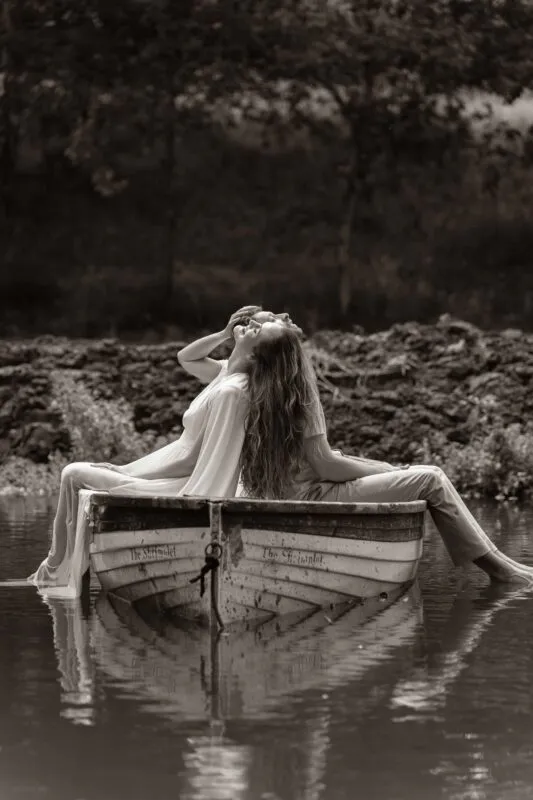A newlywed couple sits back-to-back in a small wooden rowing boat floating on a calm lake, captured in a sepia tone.