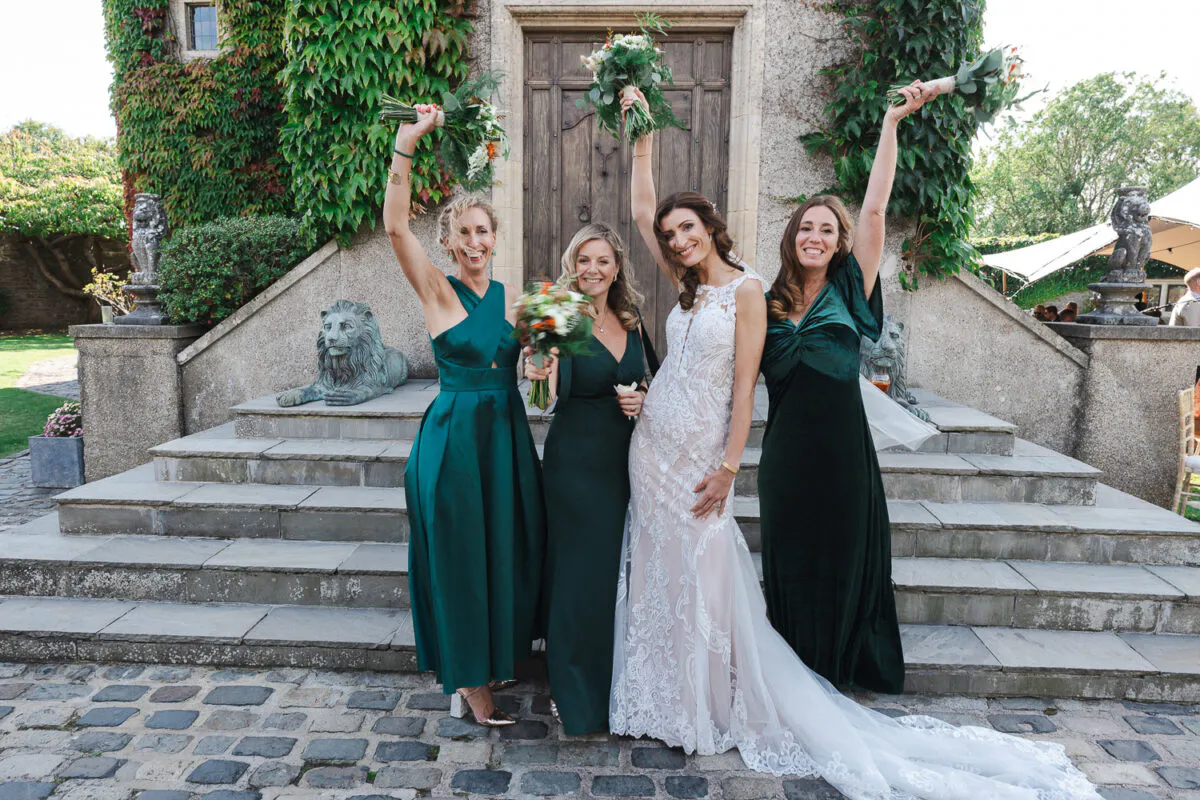 A smiling bride in a lace illusion-neckline gown poses in a cobbled courtyard with her bridesmaids, who are wearing emerald green dresses and raising their bouquets in celebration.