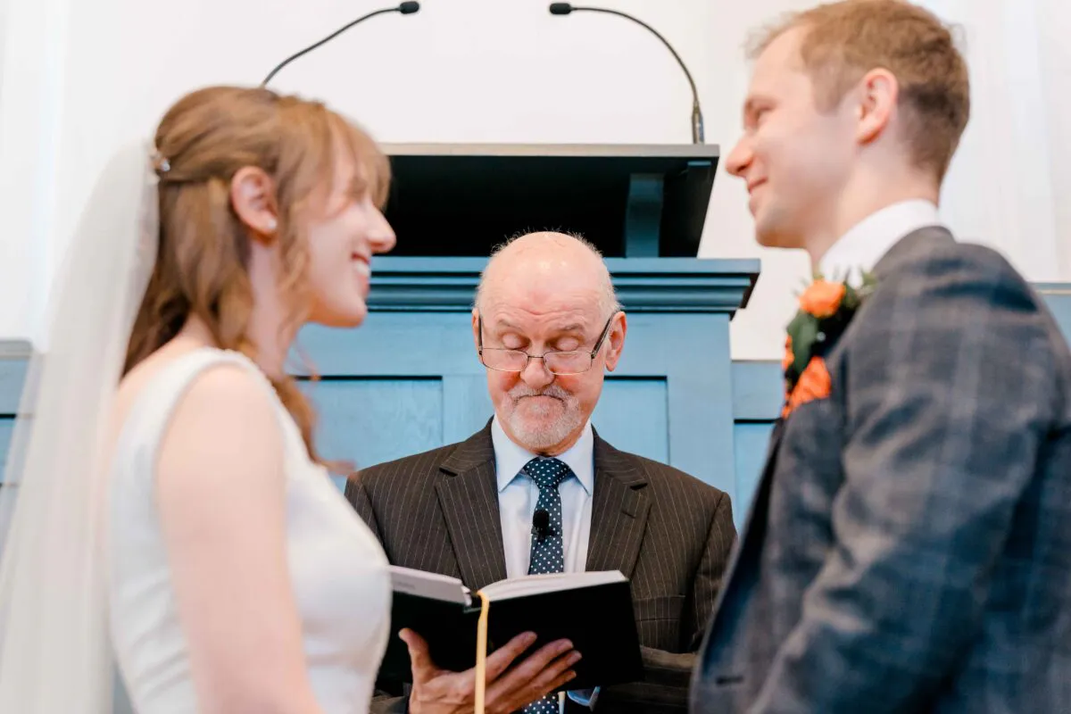 A smiling bride and groom stand facing each other during their civil ceremony while an officiant reads from a book between them.