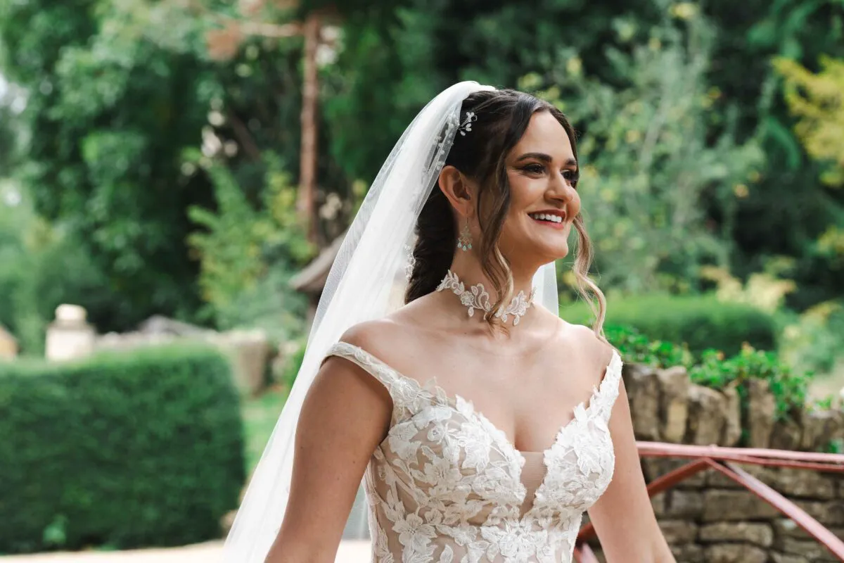 A smiling bride wearing an off-the-shoulder lace wedding dress and a fingertip veil poses in a lush green garden.