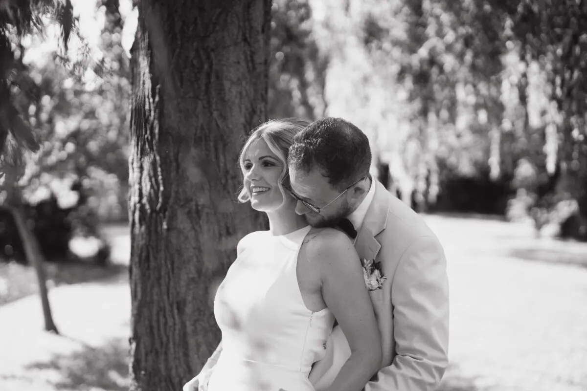 A newlywed couple shares a tender embrace in a woodland setting, captured in a monochrome documentary style with soft natural light filtering through the trees.