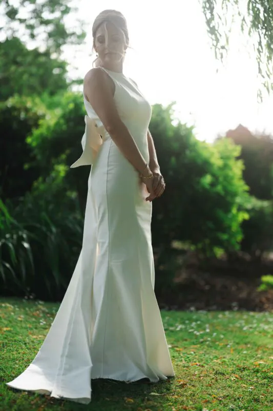 A stunning solo bride poses in a lush garden, wearing an elegant high-necked crepe column gown with a dramatic back bow and soft framing curls.