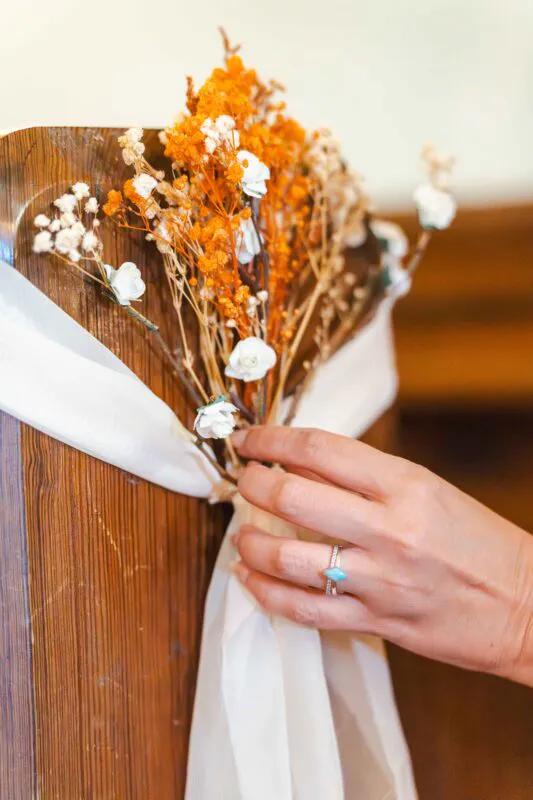 A close-up shot of a hand wearing a distinctive ring adjusting a rustic pew end decoration featuring dried orange flowers and white ribbon.