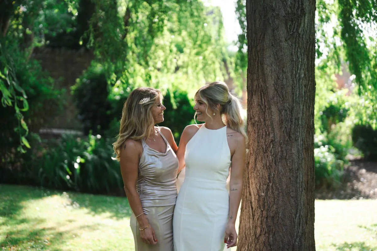 Two smiling brides stand outdoors in a sunlit garden beside a large tree, sharing a tender moment together.