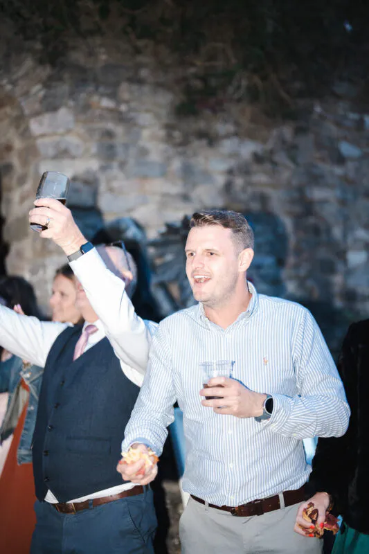A wedding guest in a striped blue shirt raises a glass in a toast while holding a small snack during an outdoor reception.