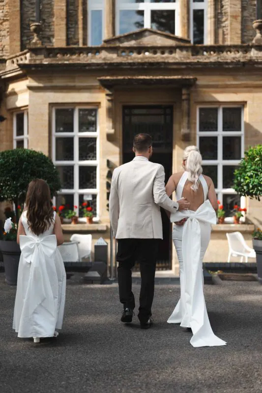 A bride, groom, and young flower girl seen from behind as they walk towards the entrance of an elegant country house hotel.