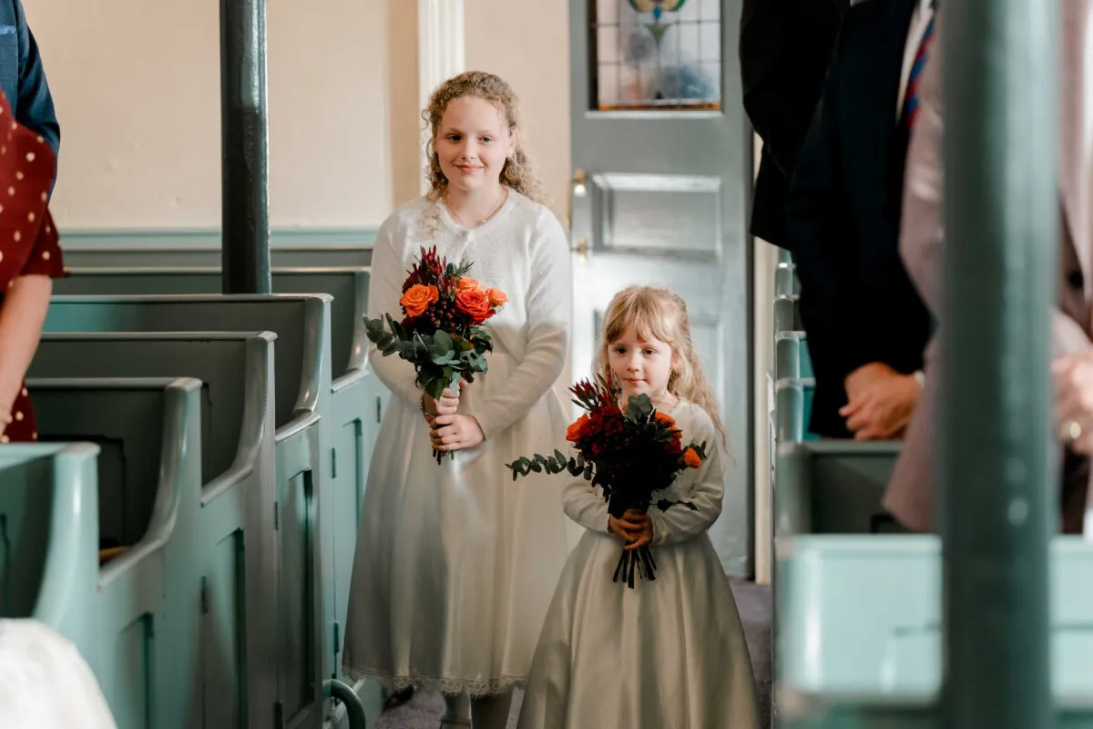 Two young flower girls in white dresses holding autumn-toned bouquets walk down the aisle between green church pews.
