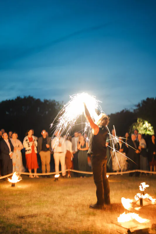 A fire performer creates a dazzling display of sparks with lit apparatuses during an evening wedding reception under a deep blue twilight sky while guests watch.