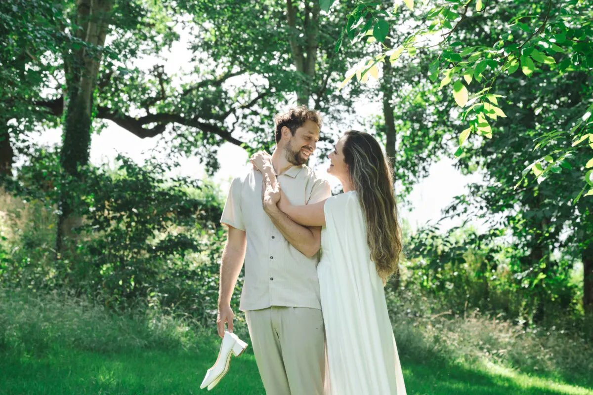 A newlywed couple smiles lovingly at each other during an outdoor portrait session in a lush green woodland surrounded by tall trees and dappled sunlight.