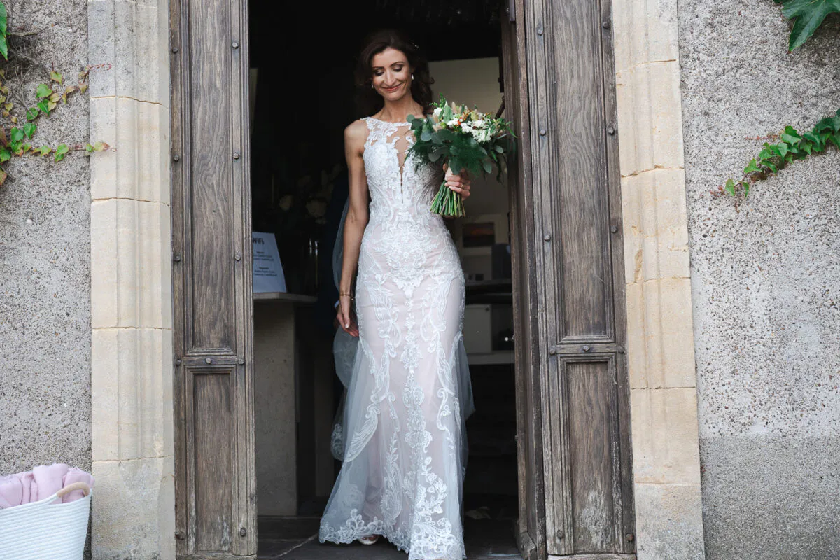 A smiling bride holding a lush greenery bouquet steps out through a grand wooden doorway framed by stone and ivy.