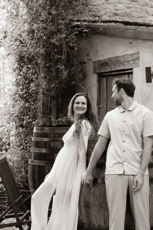 A newlywed couple holds hands and smiles at each other outside a rustic stone building with wine barrels and climbing foliage.