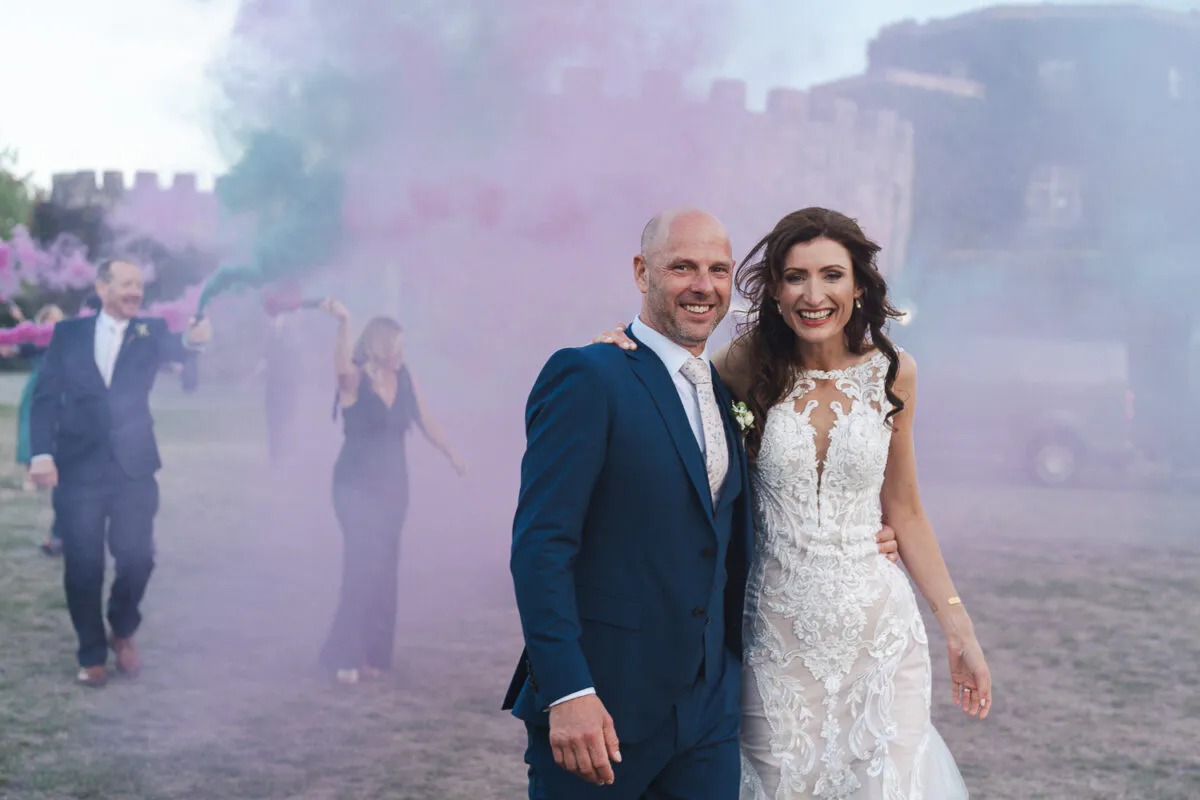 A newlywed couple smiles broadly as they walk together outdoors, framed by dramatic pink smoke flares and a distant castle backdrop.