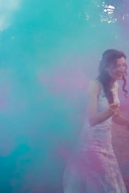 A smiling bride wearing a lace wedding dress emerges playfully through a dramatic cloud of turquoise and pink smoke.
