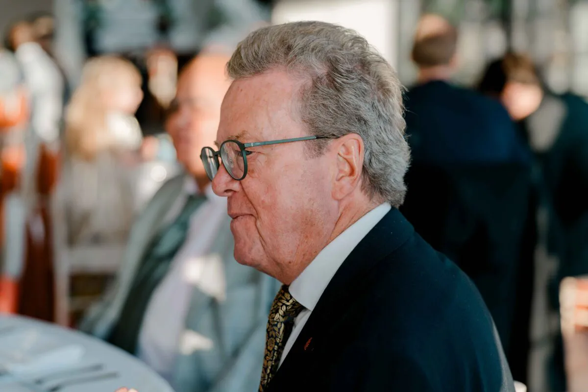 An older male guest wearing glasses, a dark suit, and a patterned tie is captured in profile during the wedding speeches.